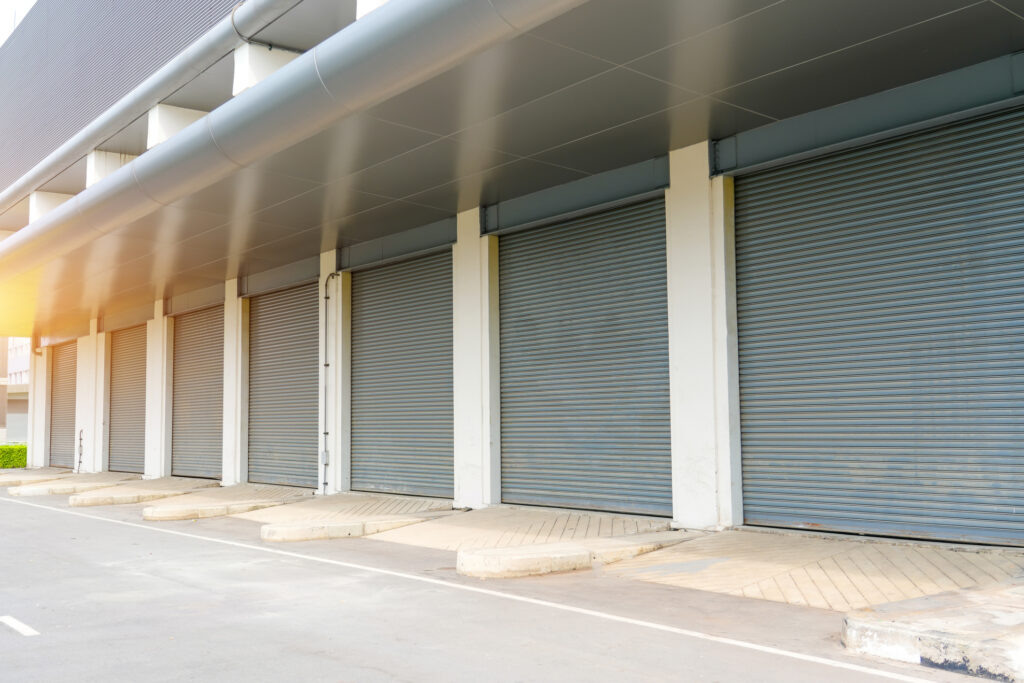 A series of metallic rolling steel doors line the exterior of a commercial building, reflecting the soft light. The parking area is empty, indicating it is early morning in a city setting.