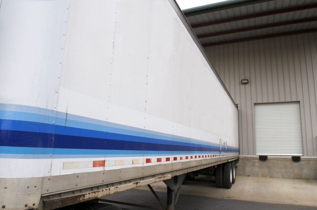 A semi-trailer loading or unloading at a small business loading dock with an empty loading bay visible with two dock bumpers next to it.