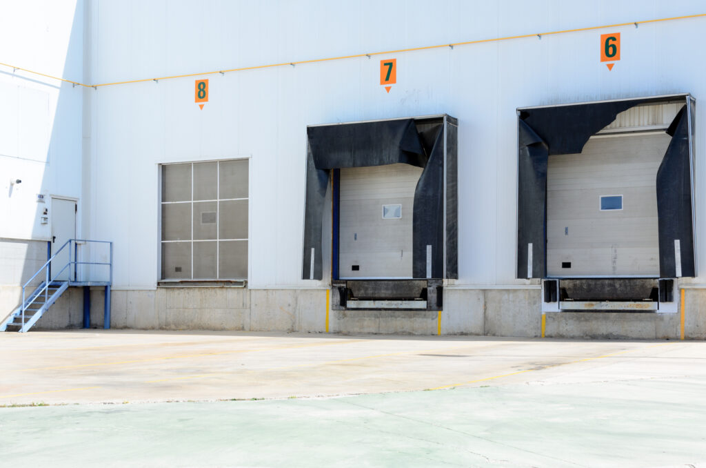 Head-on view of two empty loading dock doors of a white warehouse with one of the loading dock’s shelters is ripped off at the top.