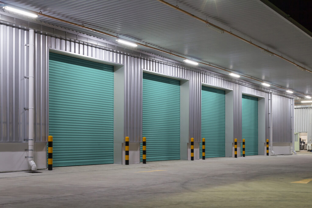Row of industrial overhead doors at a commercial loading dock, featuring rolling steel design and protective bollards under canopy lighting.