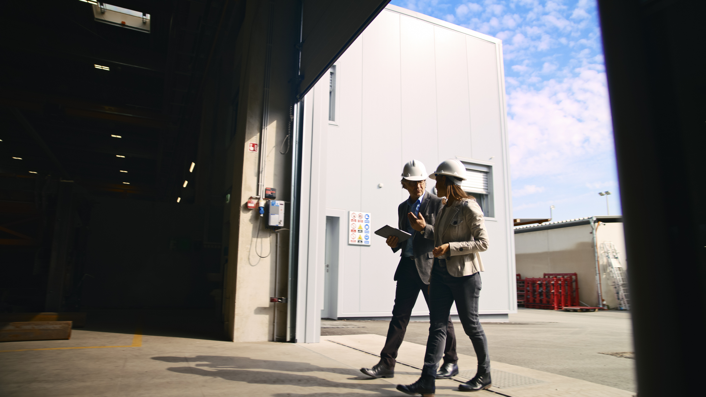 Alt text: Two professionals wearing hard hats walk outside a commercial facility near a large open overhead door, representing a trusted overhead door company on-site.